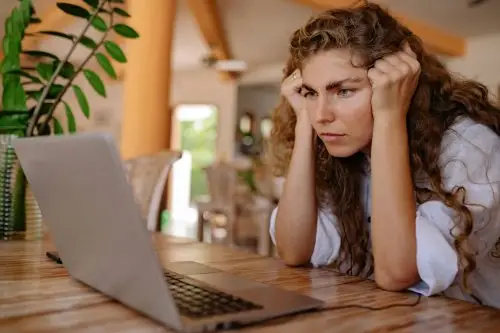 A woman with curly hair intensely focusing on her laptop indoors with plants and natural lighting.
