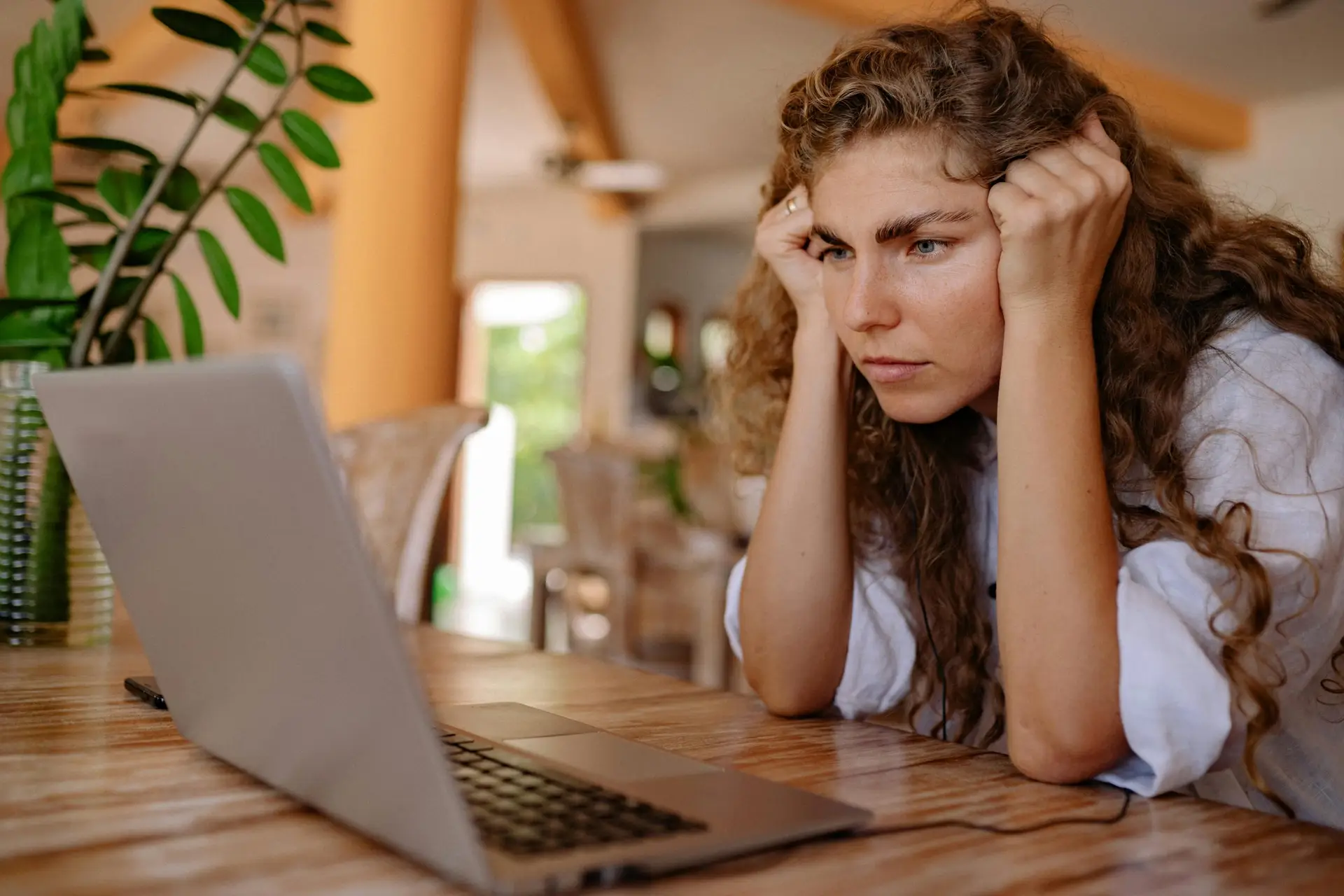 A woman with curly hair intensely focusing on her laptop indoors with plants and natural lighting.